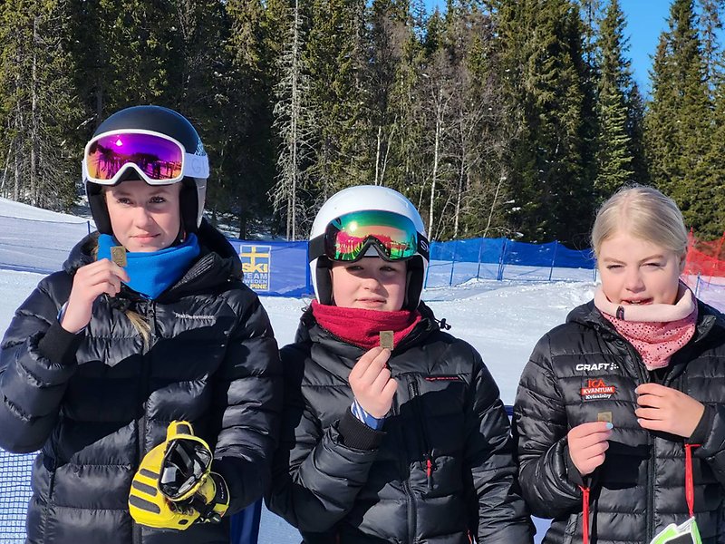 Tre tjejer som står ute i vintersol, de har vunnit lagmedalj och håller fram sina medaljer. Två av dem bär slalomhjälm.